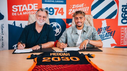 Two men sit at a table signing documents, with a bright graphic wall behind them.",
