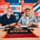 Two men sit at a table signing documents, with a bright graphic wall behind them.",