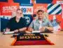 Two men sit at a table signing documents, with a bright graphic wall behind them.",
