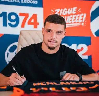 Man signing autographs at a table in front of a bright orange and blue backdrop with logos.
