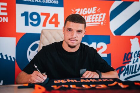 Man signing autographs at a table in front of a bright orange and blue backdrop with logos.
