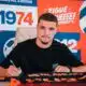 Man signing autographs at a table in front of a bright orange and blue backdrop with logos.