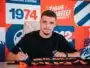Man signing autographs at a table in front of a bright orange and blue backdrop with logos.