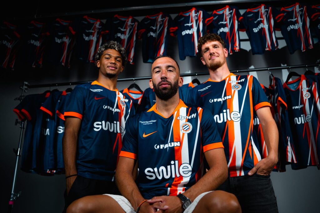 Three male athletes in blue and orange striped jerseys pose together in a locker room, with team uniforms hanging on racks behind them.