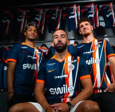 Three male athletes in blue and orange striped jerseys pose together in a locker room, with team uniforms hanging on racks behind them.