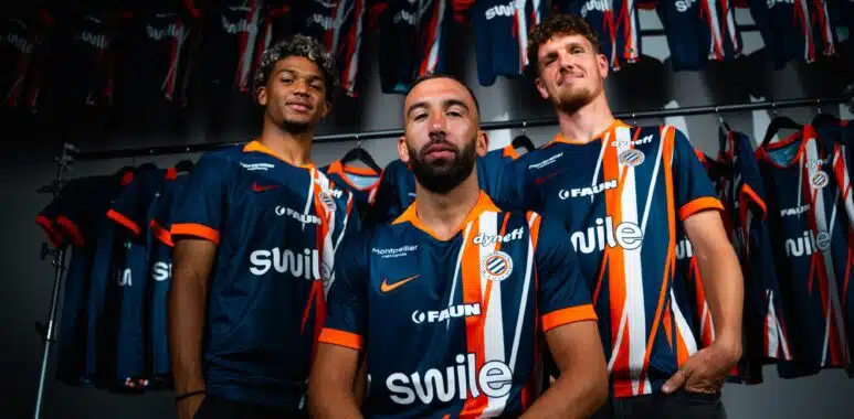 dsc01439-2048x1365 | AllezPaillade.com Three male athletes in blue and orange striped jerseys pose together in a locker room, with team uniforms hanging on racks behind them.