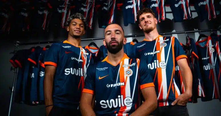 dsc01439-2048x1365 | AllezPaillade.com Three male athletes in blue and orange striped jerseys pose together in a locker room, with team uniforms hanging on racks behind them.