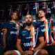 Three male athletes in blue and orange striped jerseys pose together in a locker room, with team uniforms hanging on racks behind them.