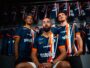 Three male athletes in blue and orange striped jerseys pose together in a locker room, with team uniforms hanging on racks behind them.