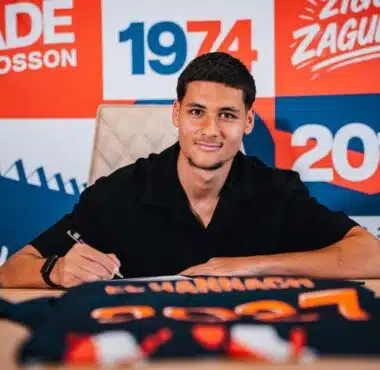 Young man signs a document at a table, with a bright red and blue graphic backdrop behind him.