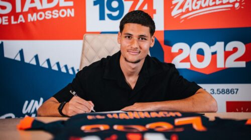 Young man signs a document at a table, with a bright red and blue graphic backdrop behind him.