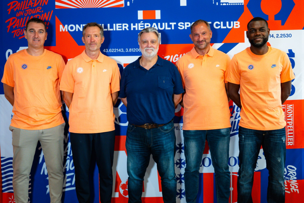 image | AllezPaillade.com Five men pose together indoors, four in orange polo shirts and one in a navy shirt, standing in front of a blue/orange graphic backdrop with text and logos.