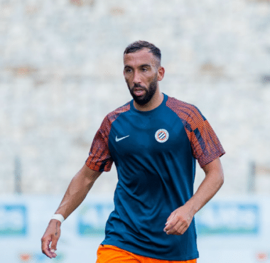 Male soccer player in a blue training shirt and orange shorts on a blurred stadium field, mid-action pose.