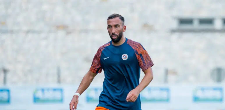 image | AllezPaillade.com Male soccer player in a blue training shirt and orange shorts on a blurred stadium field, mid-action pose.