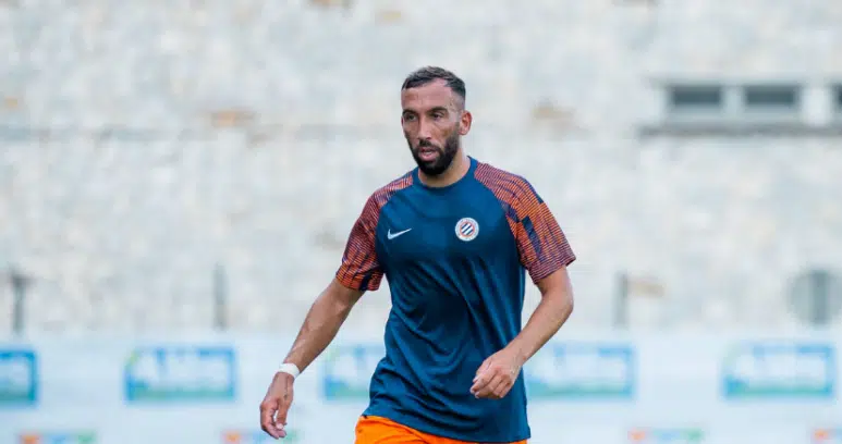 image | AllezPaillade.com Male soccer player in a blue training shirt and orange shorts on a blurred stadium field, mid-action pose.