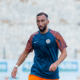 Male soccer player in a blue training shirt and orange shorts on a blurred stadium field, mid-action pose.