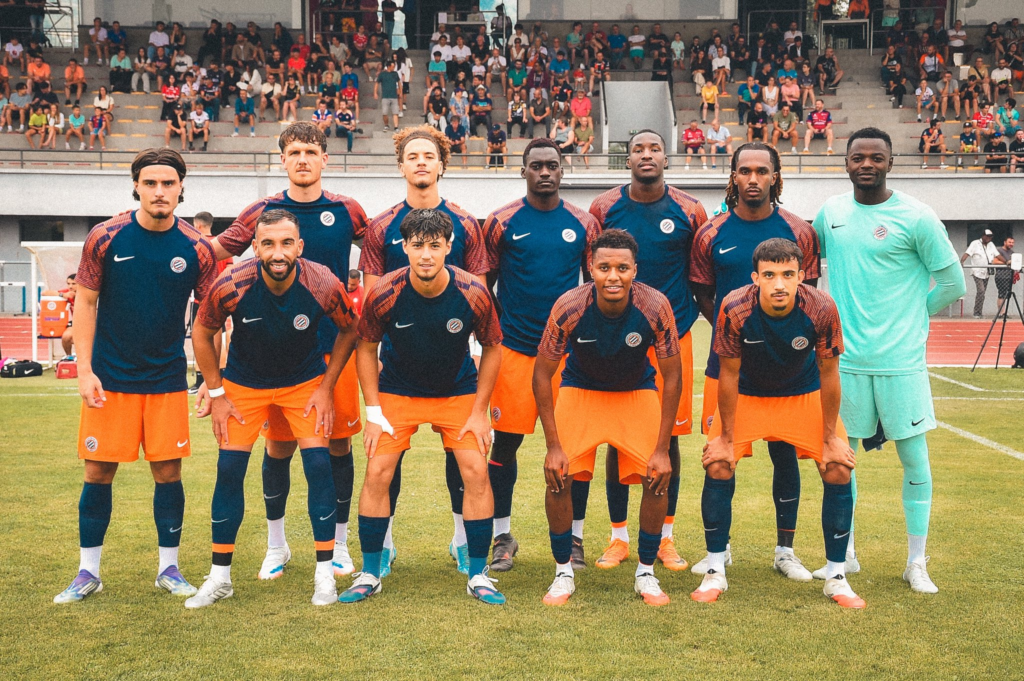 Soccer team in blue and orange kits poses for a group photo on the field, with spectators in the stands behind them.