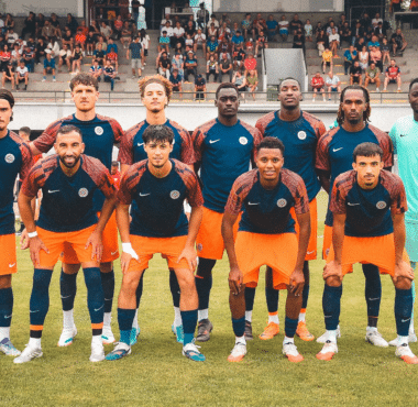 Soccer team in blue and orange kits poses for a group photo on the field, with spectators in the stands behind them.