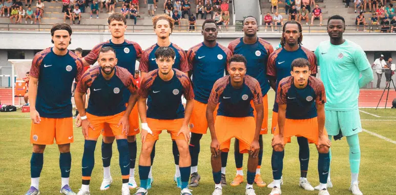 Soccer team in blue and orange kits poses for a group photo on the field, with spectators in the stands behind them.