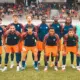 Soccer team in blue and orange kits poses for a group photo on the field, with spectators in the stands behind them.
