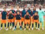 Soccer team in blue and orange kits poses for a group photo on the field, with spectators in the stands behind them.