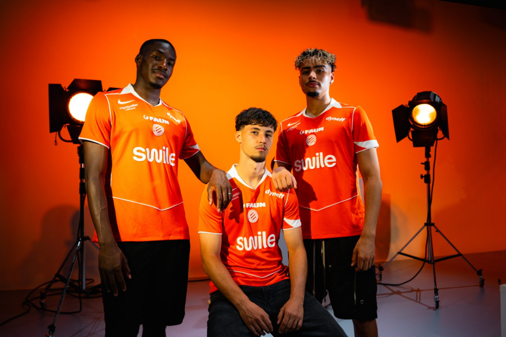 image | AllezPaillade.com Four men in bright orange sports jerseys pose for a studio team photo against an orange backdrop with lighting equipment visible behind them.