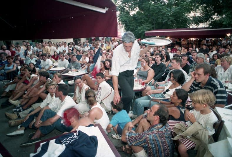 JMMAR20180606 | AllezPaillade.com Street performer balances a tray overhead as a large crowd watches at an outdoor cafe or plaza party in daylight.