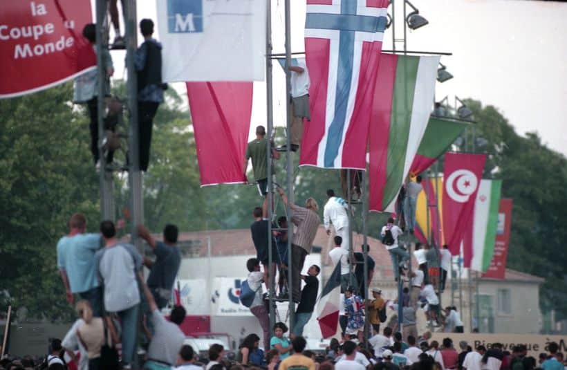 JMMAR20180606 | AllezPaillade.com Crowd at an outdoor festival with tall flagpoles displaying many international flags and people climbing the poles to adjust them.