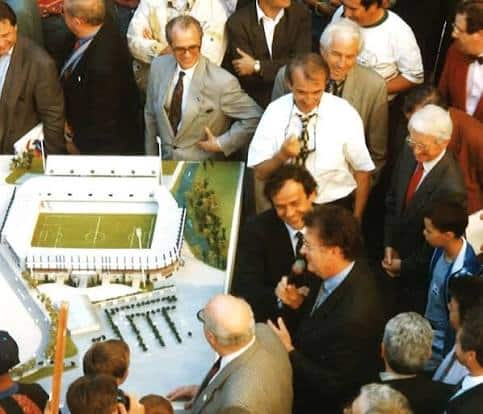 images 18 | AllezPaillade.com Crowd of men in suits gathers around a large architectural model of a soccer stadium, examining the field and stands.