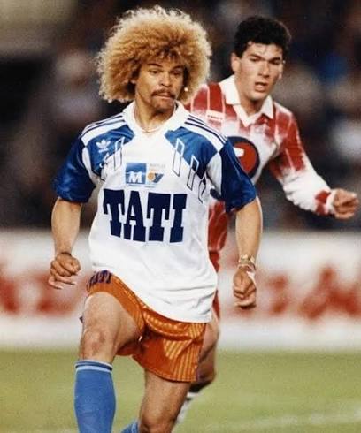 images 4 | AllezPaillade.com Soccer players sprinting on a field; foreground player with a curly blond Afro wearing a blue-white jersey labeled 'TATI' and orange shorts, pursued by a red-and-white kit opponent.
