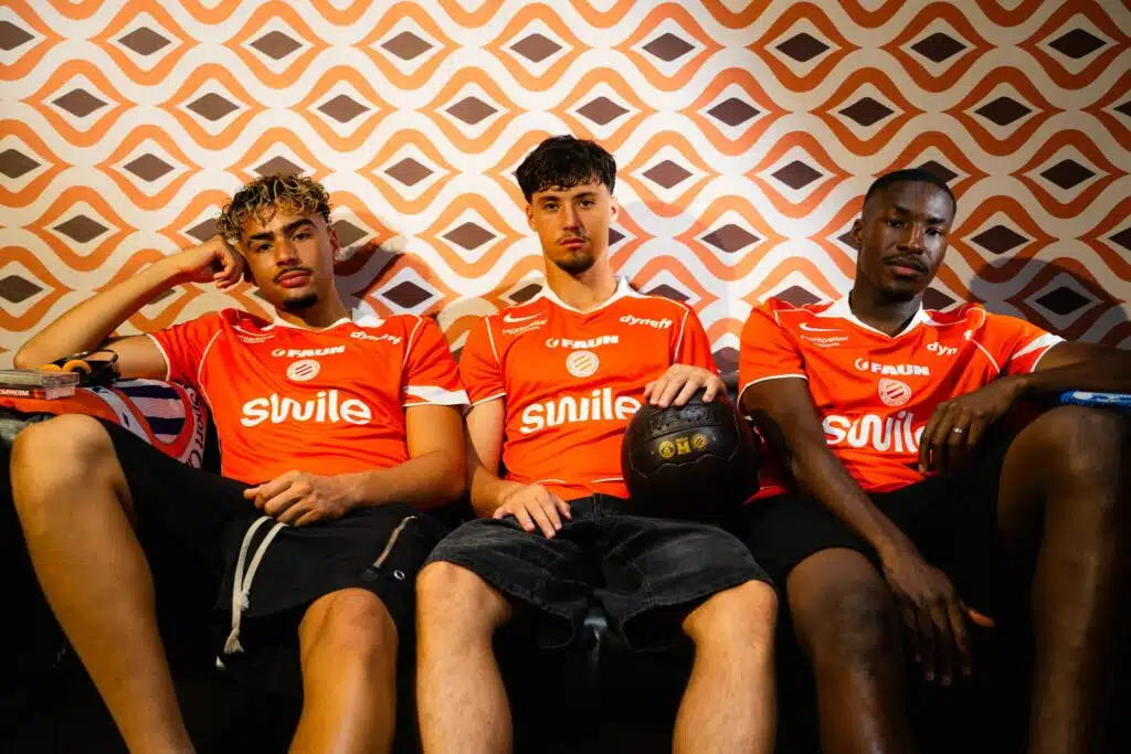 maillot | AllezPaillade.com Three young men in orange sports jerseys sit on a couch against an orange geometric wallpaper; one holds a black basketball in his lap.