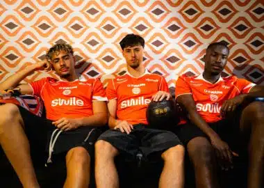 maillot | AllezPaillade.com Three young men in orange sports jerseys sit on a couch against an orange geometric wallpaper; one holds a black basketball in his lap.