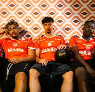 Three young men in orange sports jerseys sit on a couch against an orange geometric wallpaper; one holds a black basketball in his lap.