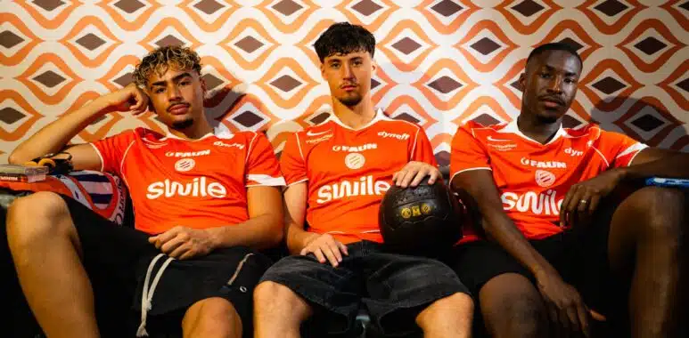 maillot | AllezPaillade.com Three young men in orange sports jerseys sit on a couch against an orange geometric wallpaper; one holds a black basketball in his lap.