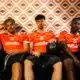 Three young men in orange sports jerseys sit on a couch against an orange geometric wallpaper; one holds a black basketball in his lap.
