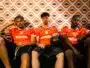Three young men in orange sports jerseys sit on a couch against an orange geometric wallpaper; one holds a black basketball in his lap.