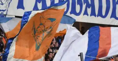 Crowd of sports fans waving orange, blue, and white flags in a stadium with a large blue-letter banner above them.