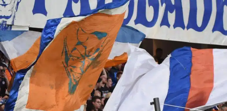 montpellier-la-mosson | AllezPaillade.com Crowd of sports fans waving orange, blue, and white flags in a stadium with a large blue-letter banner above them.