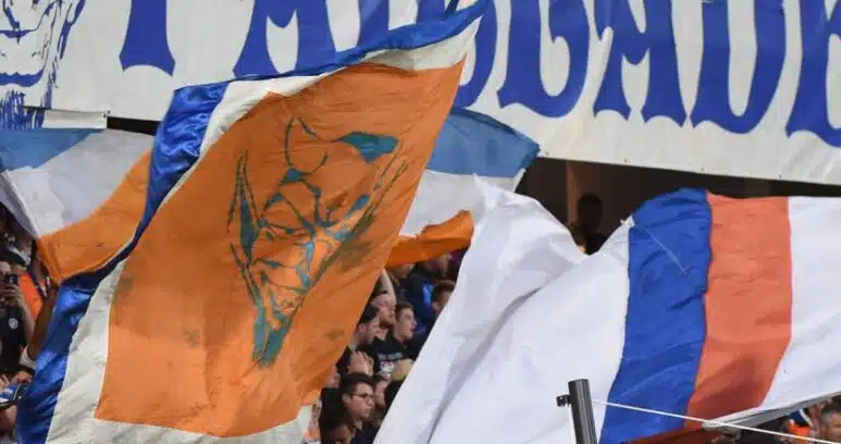 Crowd of sports fans waving orange, blue, and white flags in a stadium with a large blue-letter banner above them.
