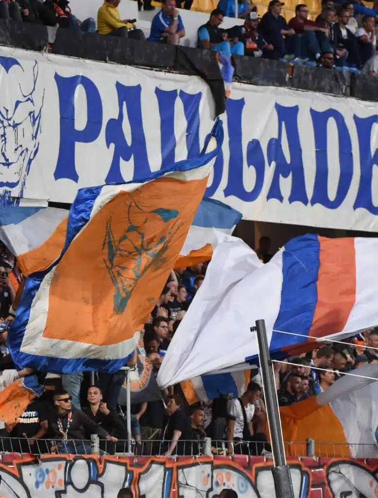 montpellier-la-mosson | AllezPaillade.com Crowd of sports fans waving orange, blue, and white flags in a stadium with a large blue-letter banner above them.
