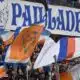 Crowd of sports fans waving orange, blue, and white flags in a stadium with a large blue-letter banner above them.