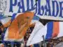 Crowd of sports fans waving orange, blue, and white flags in a stadium with a large blue-letter banner above them.