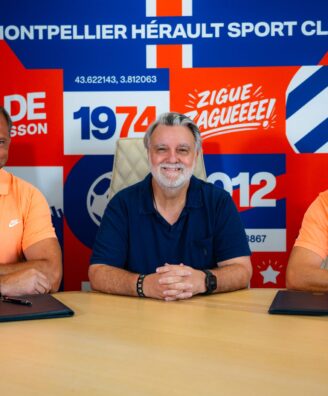 Three men sit at a table in front of a bright, graphic backdrop; left and right wear orange polo shirts while the center wears a dark blue shirt, posing for a team photo in front of a sports-club banner.