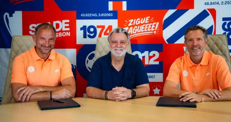 Three men sit at a table in front of a bright, graphic backdrop; left and right wear orange polo shirts while the center wears a dark blue shirt, posing for a team photo in front of a sports-club banner.