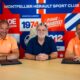 Three men sit at a table in front of a bright, graphic backdrop; left and right wear orange polo shirts while the center wears a dark blue shirt, posing for a team photo in front of a sports-club banner.