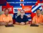 Three men sit at a table in front of a bright, graphic backdrop; left and right wear orange polo shirts while the center wears a dark blue shirt, posing for a team photo in front of a sports-club banner.