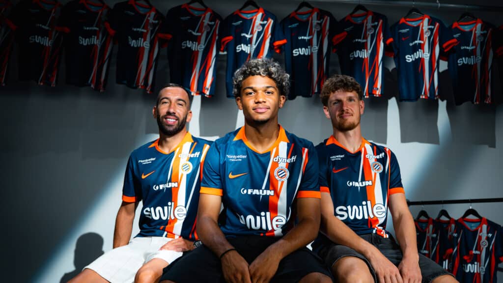 nouveau-maillot-mise-en-avant | AllezPaillade.com Three soccer players in blue-orange striped jerseys sit together in a locker room with team jerseys hanging in the background.