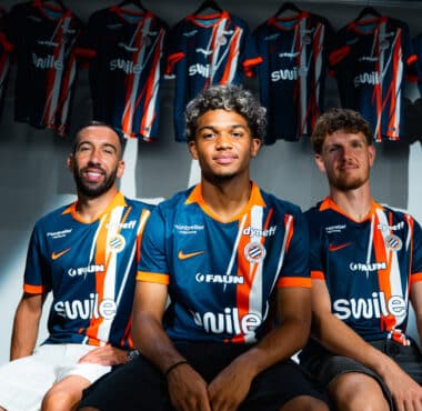 Three soccer players in blue-orange striped jerseys sit together in a locker room with team jerseys hanging in the background.