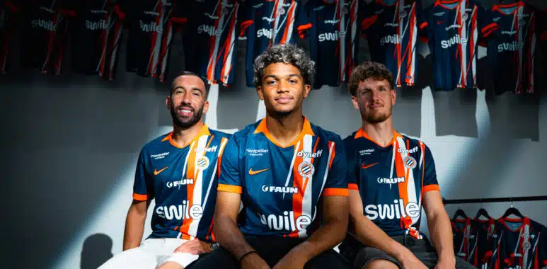 Three soccer players in blue-orange striped jerseys sit together in a locker room with team jerseys hanging in the background.
