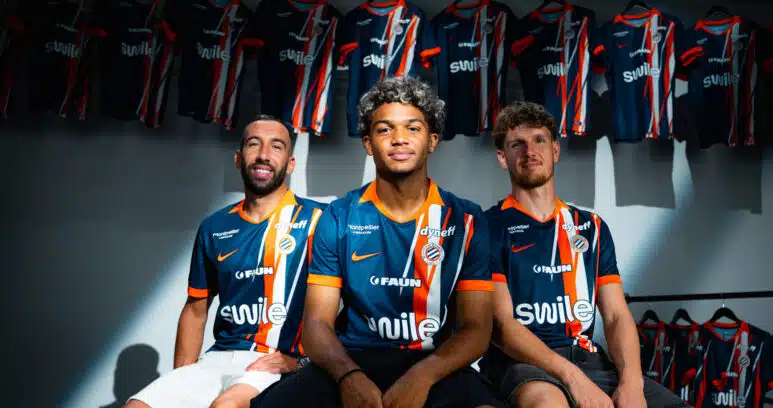 nouveau-maillot-mise-en-avant | AllezPaillade.com Three soccer players in blue-orange striped jerseys sit together in a locker room with team jerseys hanging in the background.